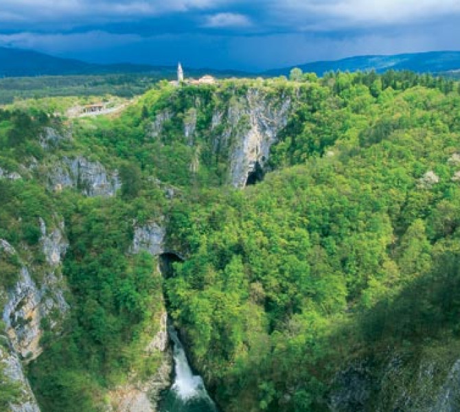 Forest-covered rocks; a river flows out of the cave.