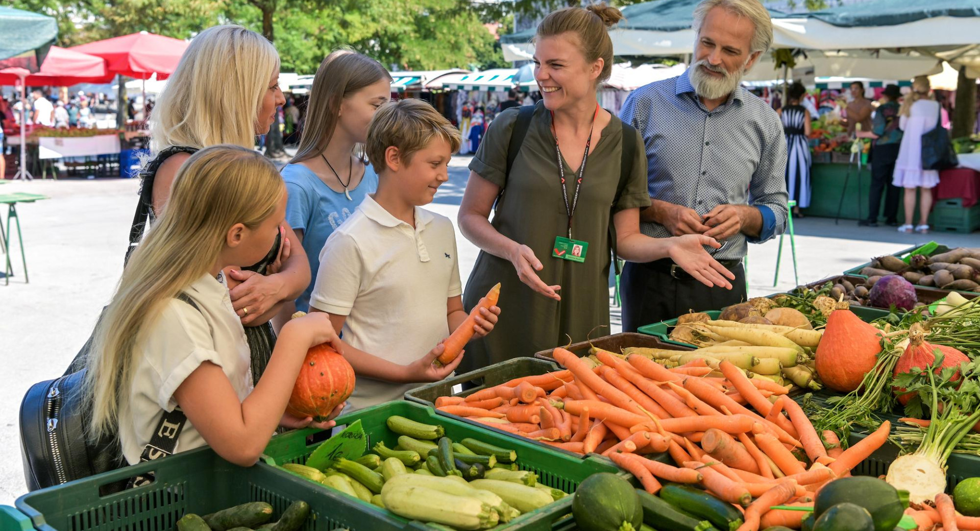 Druzina z vodnico na trznici si ogleduje ponudbo zelenjave.