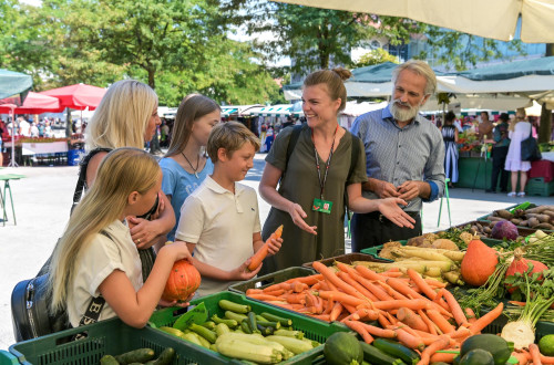 Druzina z vodnico na trznici si ogleduje ponudbo zelenjave.