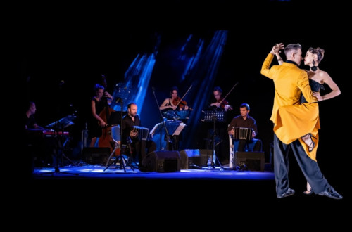 In the foreground, on the right, an illuminated couple dances in an embrace; in the background, on the left, musicians sit and play instruments in the dark under blue spotlights.