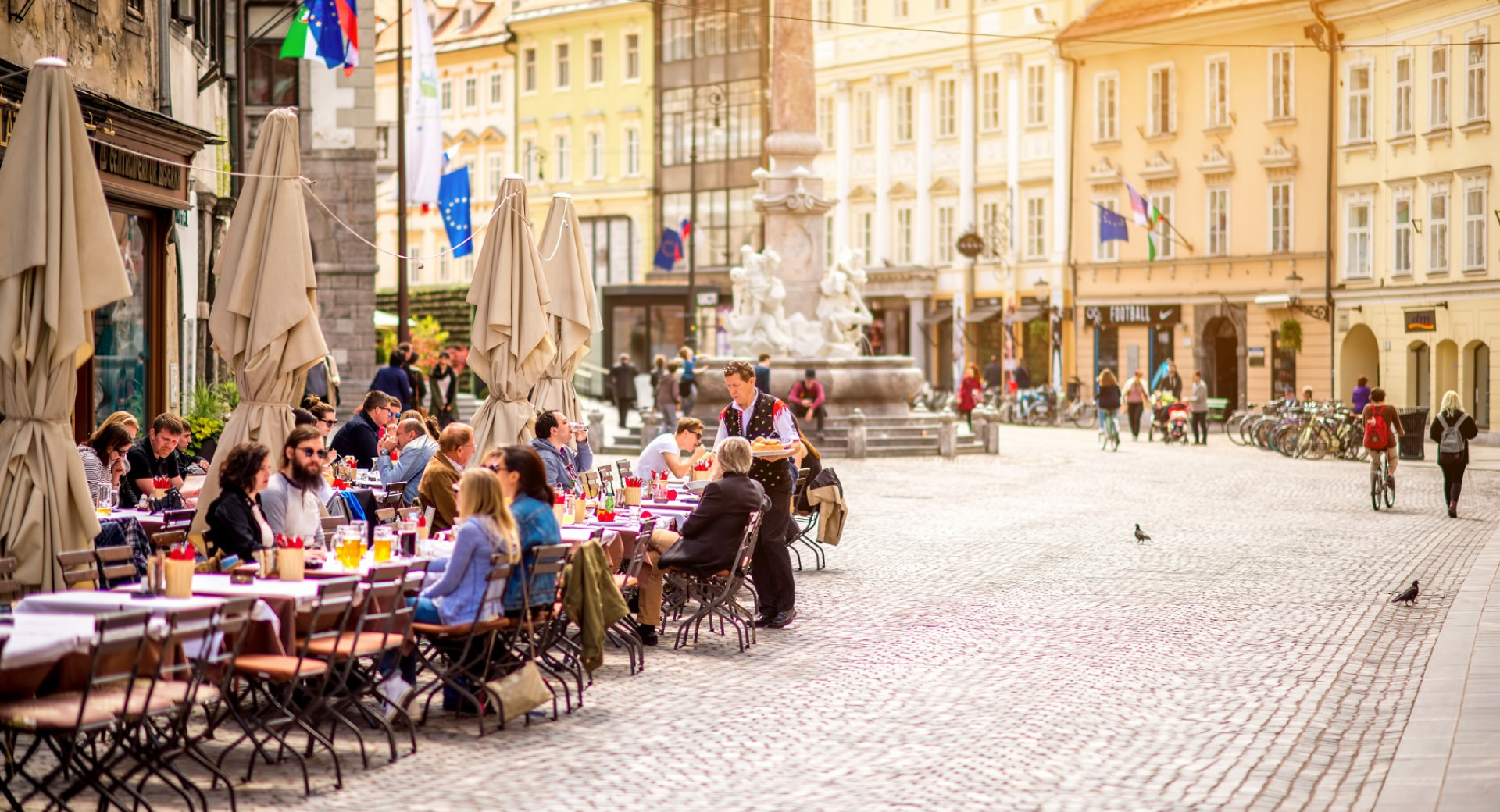 Ljudje sedijo na soncu v kavarni v starem mestnem jedru. V ozadju stare rumene mescanske stavbe. V ospredju ljudje ob kavi.