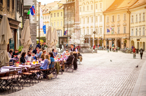 People sitting in the sun in a cafe on a city square. Drinking coffee. Old yellow buildings in the background.