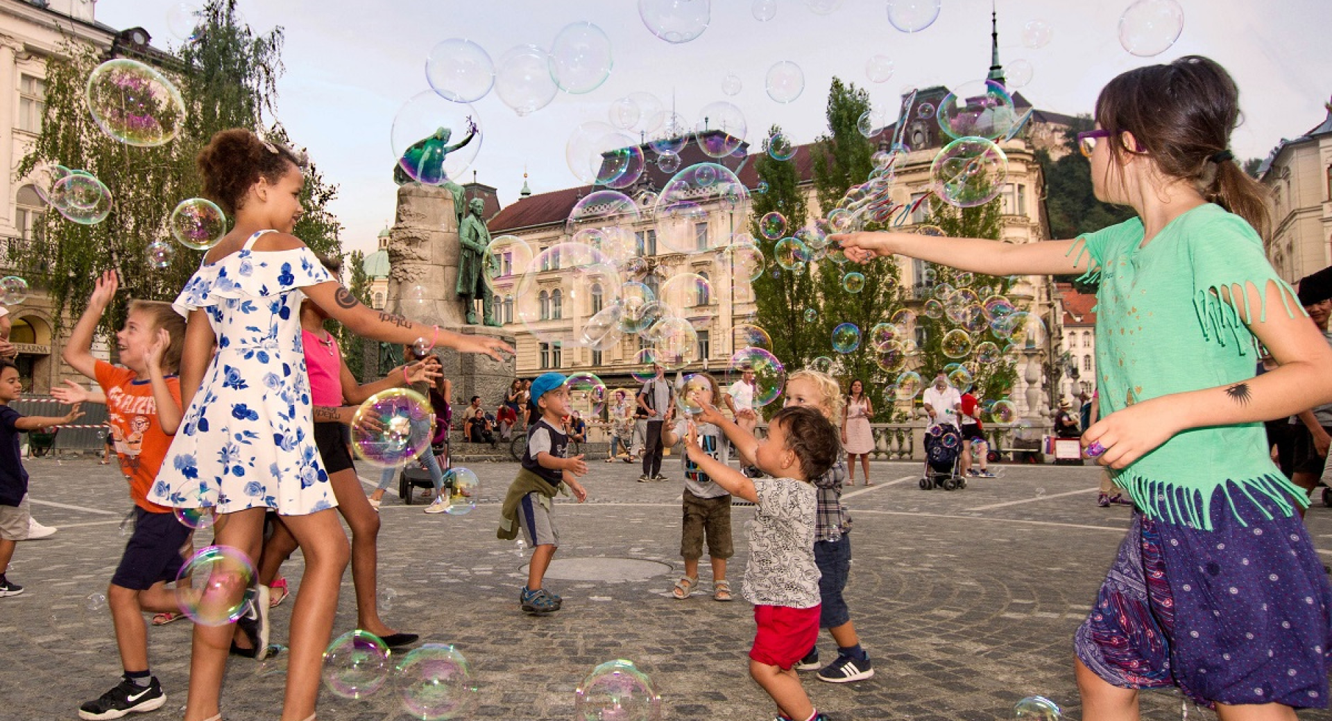 Children of different age are catching bubbles in the city square.
