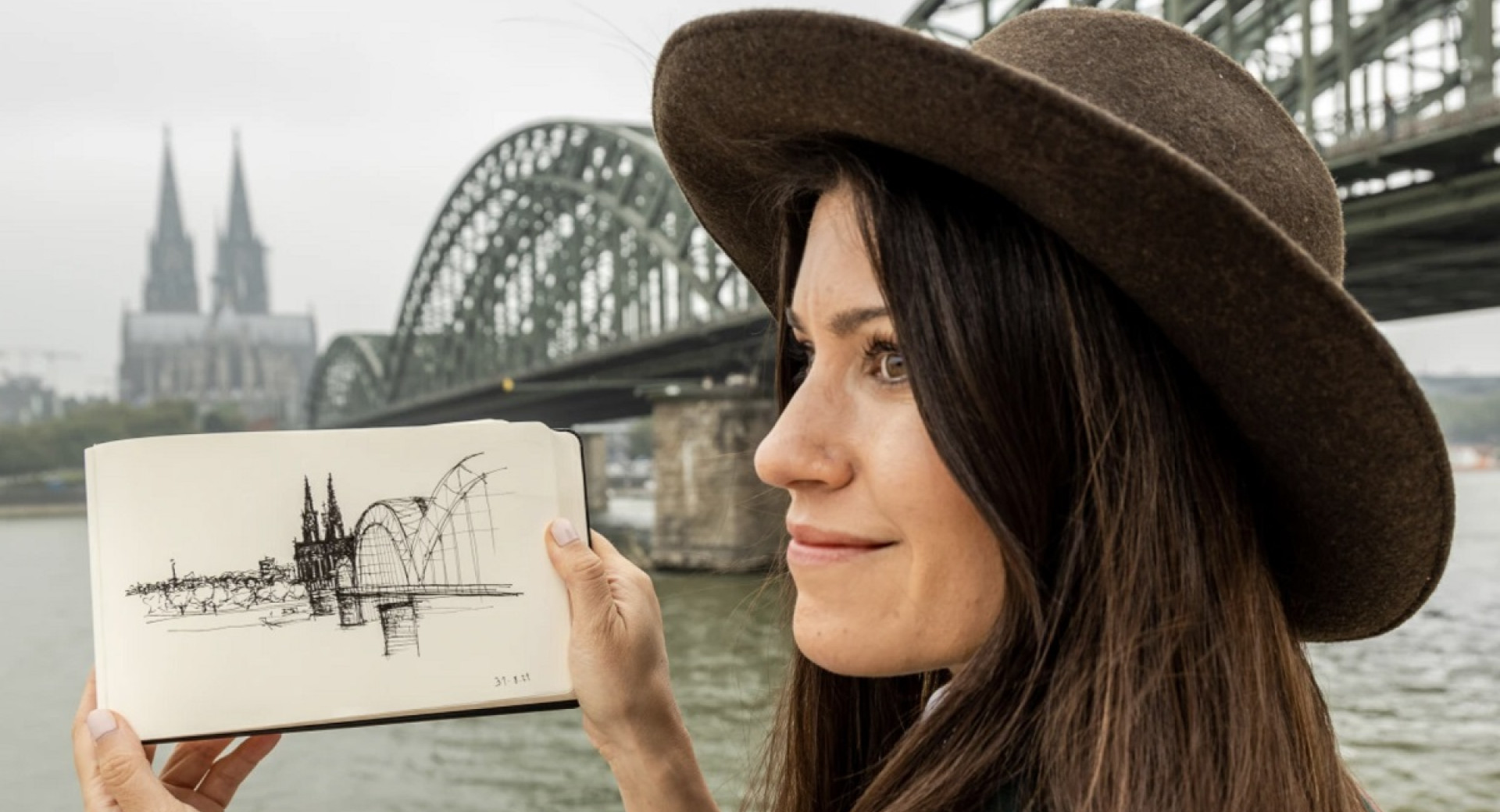 A profile photograph of a young woman holding a sketch. A large steel bridge in the background.