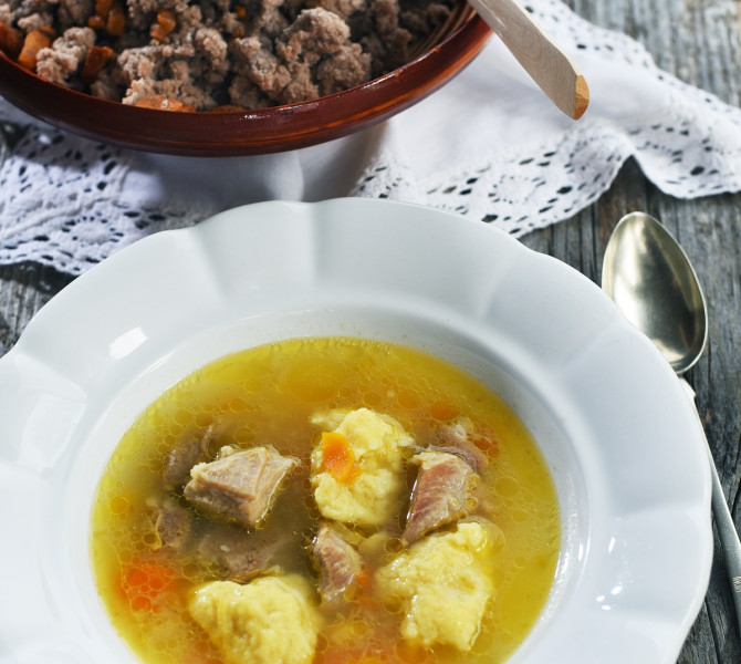 Soup with meat and carrots in a white plate in the foreground. Buckwheat žganci in the background.