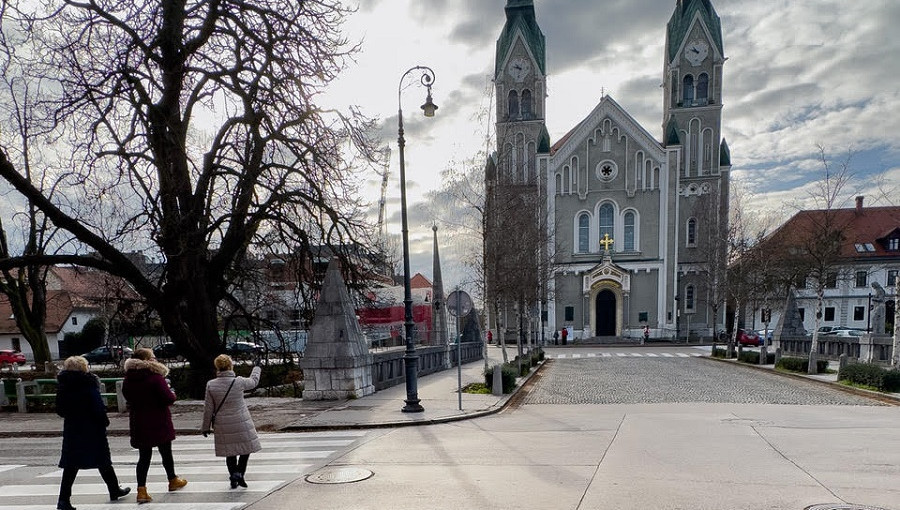 Velika siva cerkev z dvema zvonikoma. Pred njo most.