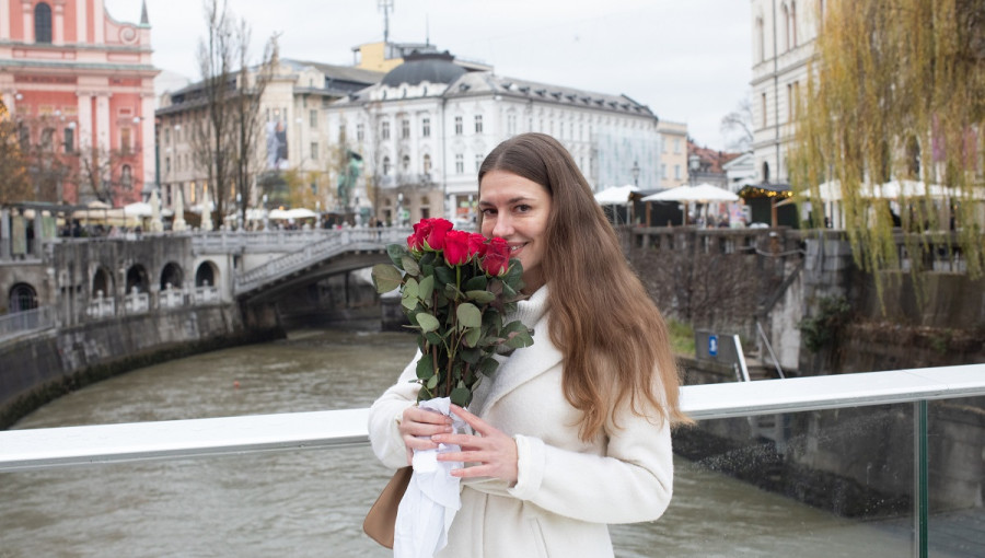 A young woman with a bouque of red roses standing on a glass bridge across a river. A beautiful cityscape in the background.