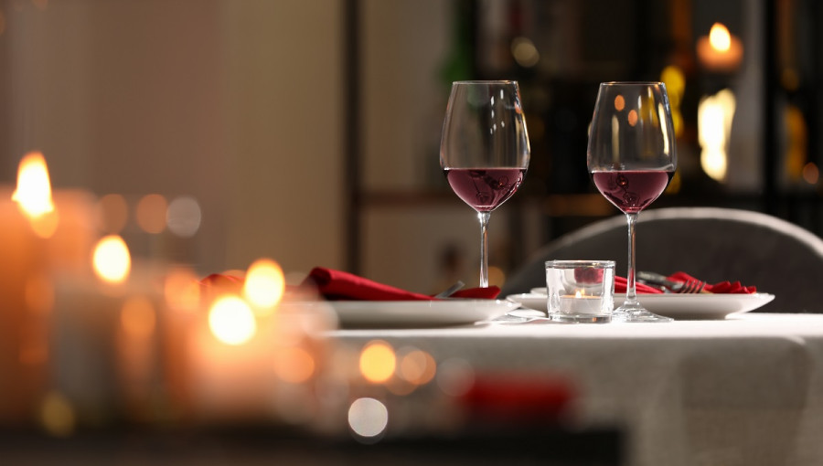 A table with white cloth. Two glasses of red wine on it. Lit candles in the foreground.