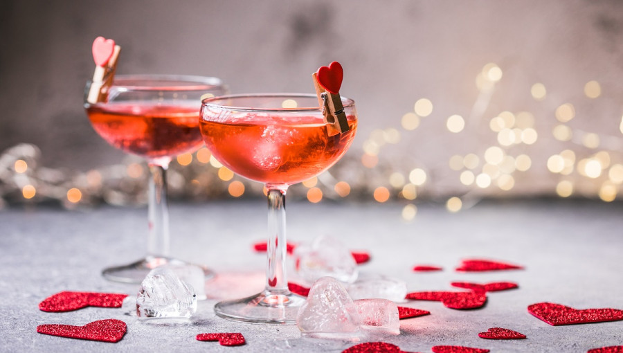 Two glasses with cocktail on a grey surface. Red hearts scattered around them.
