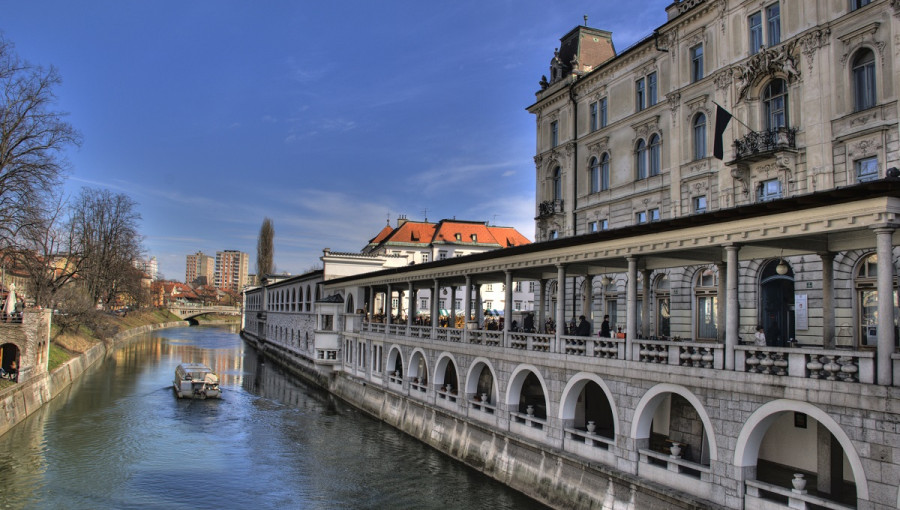 A river boat in the middle of a river in the city. Wonderful arcades next to it belonging to a historical building.