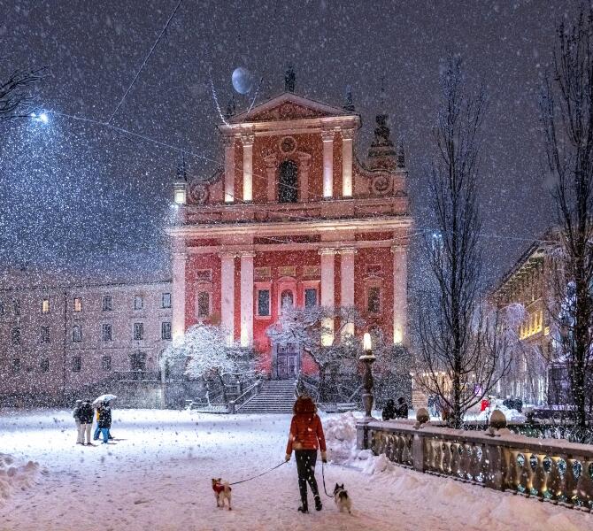 A snowy scene at Prešeren Square with the Franciscan Church in the background, a person walking dogs in the foreground.