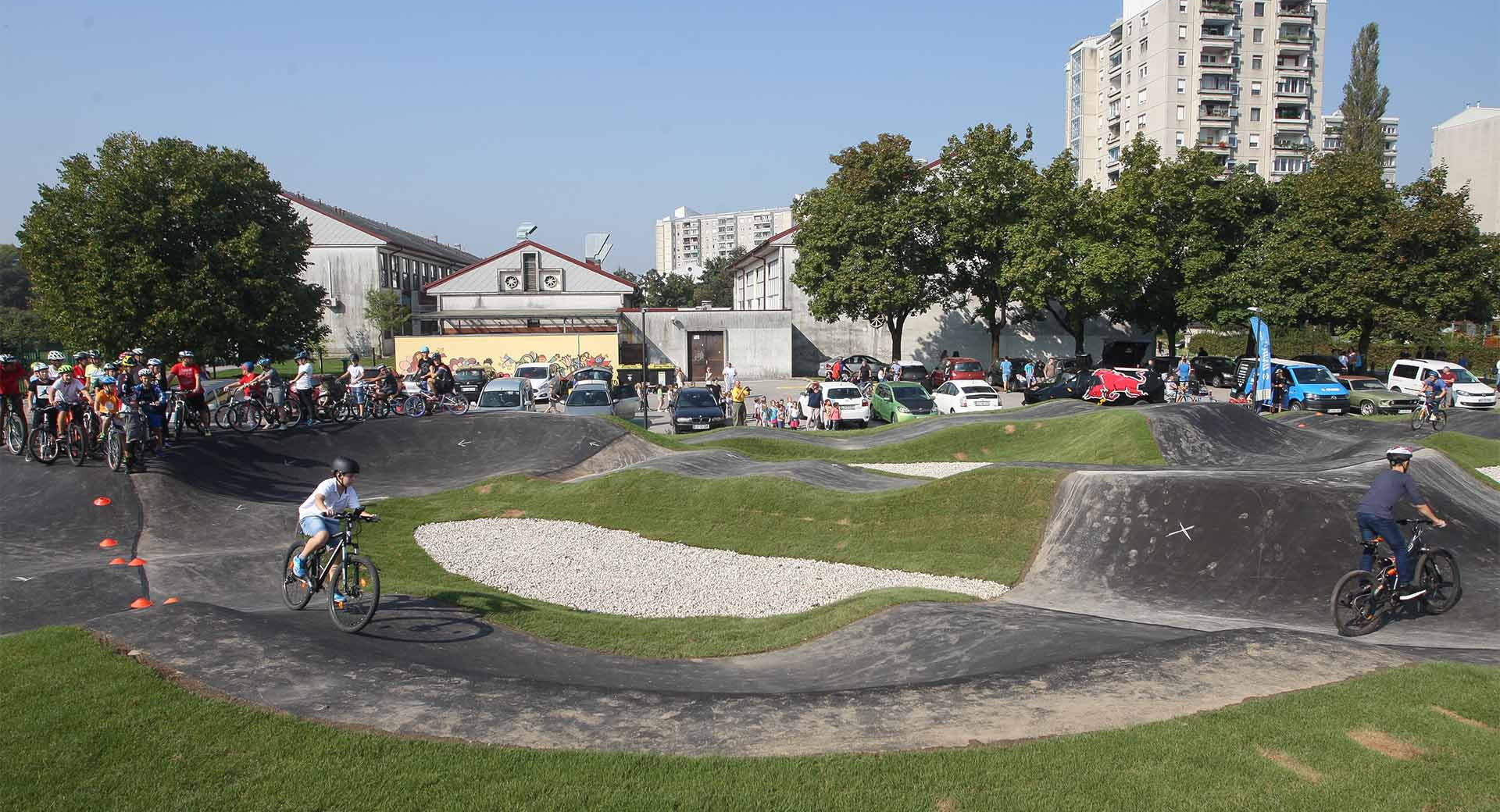 Otroci in mladostniki na pumptrack poligonu vozijo kolesa po grbinah med mnozico gledalcev na soncnem mestnem dogodku.