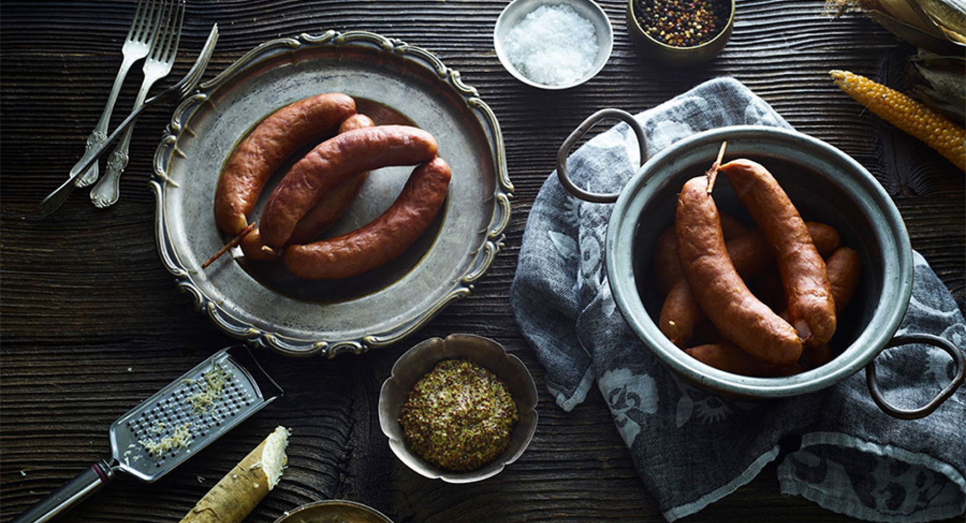 Smoked sausages are arranged on a decorative plate, surrounded by a grater, spices, and salt.