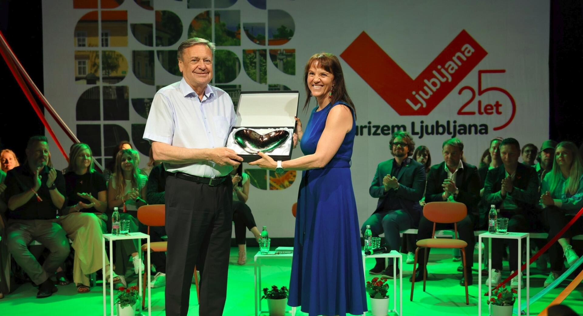 Dve osebi stojita na odru, v rokah držita priznanje v obliki srca, za njima sedijo drugi ljudje.