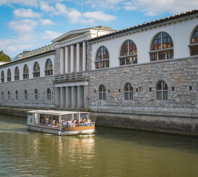 A tourist boat with passengers cruises along the Ljubljanica River past the monumental stone building of Plečnik's Market.