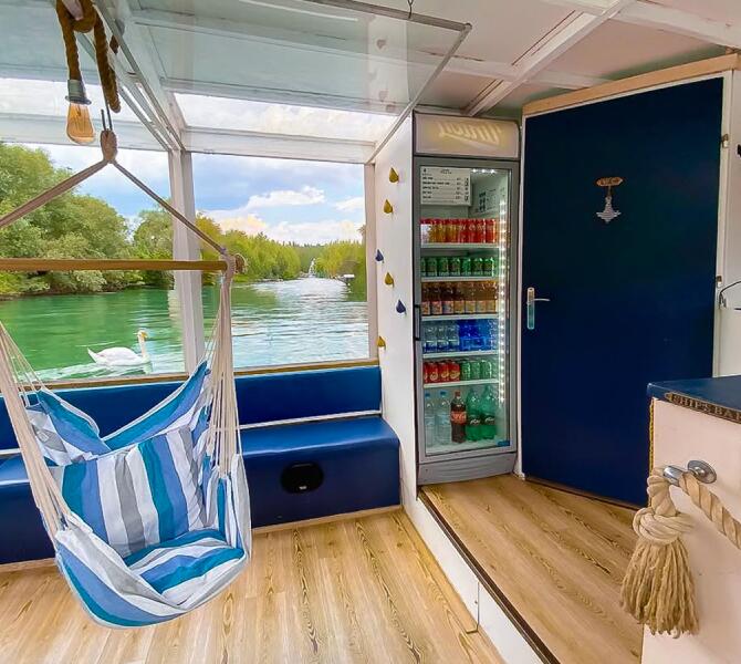The interior of the Barka Lea boat featuring a hanging chair, a drinks fridge, and a window view of a swan and green riverbanks.