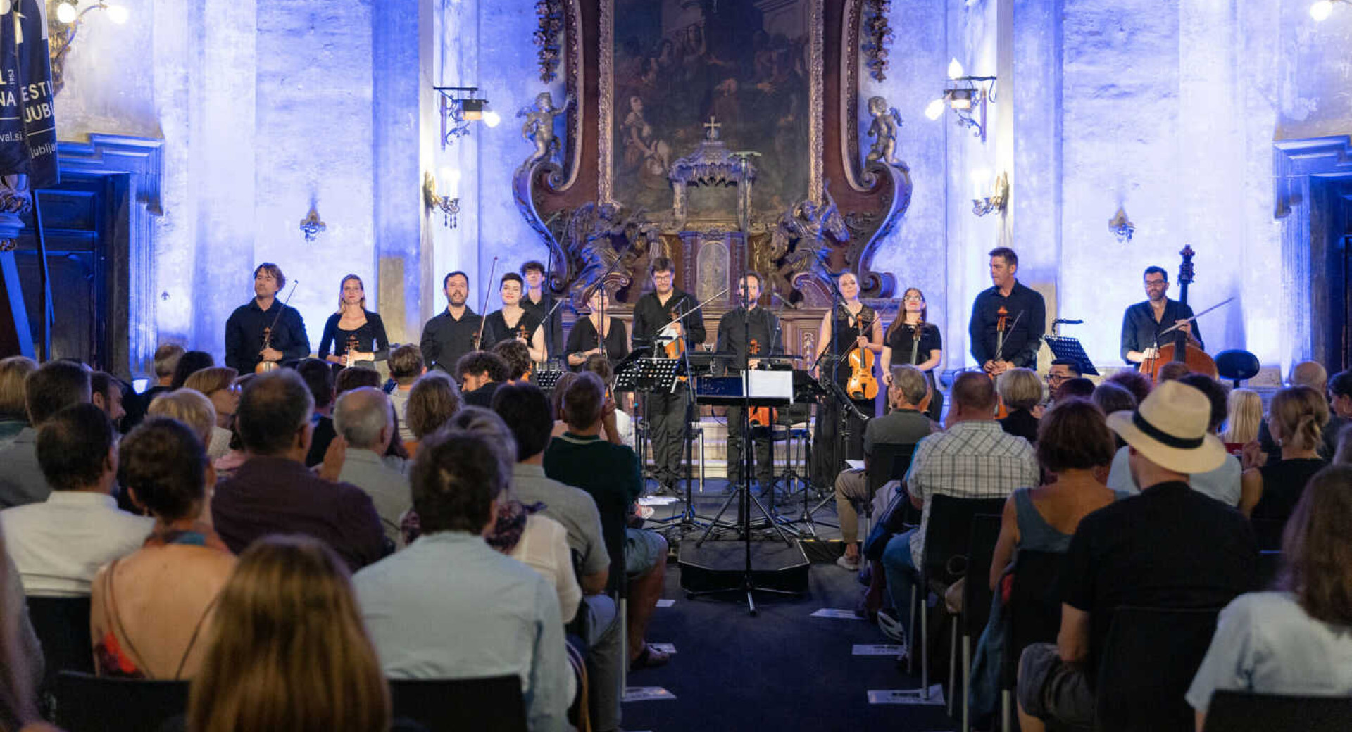 Zbor poje v cerkvi, gledalci pa opazujejo; za njimi je ozadje osvetljeno z belo-modro svetlobo.