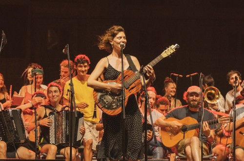 Etno orkester sedi in igra na instrumente, pred njimi pa stoji pevka s kitaro in poje v mikrofon.