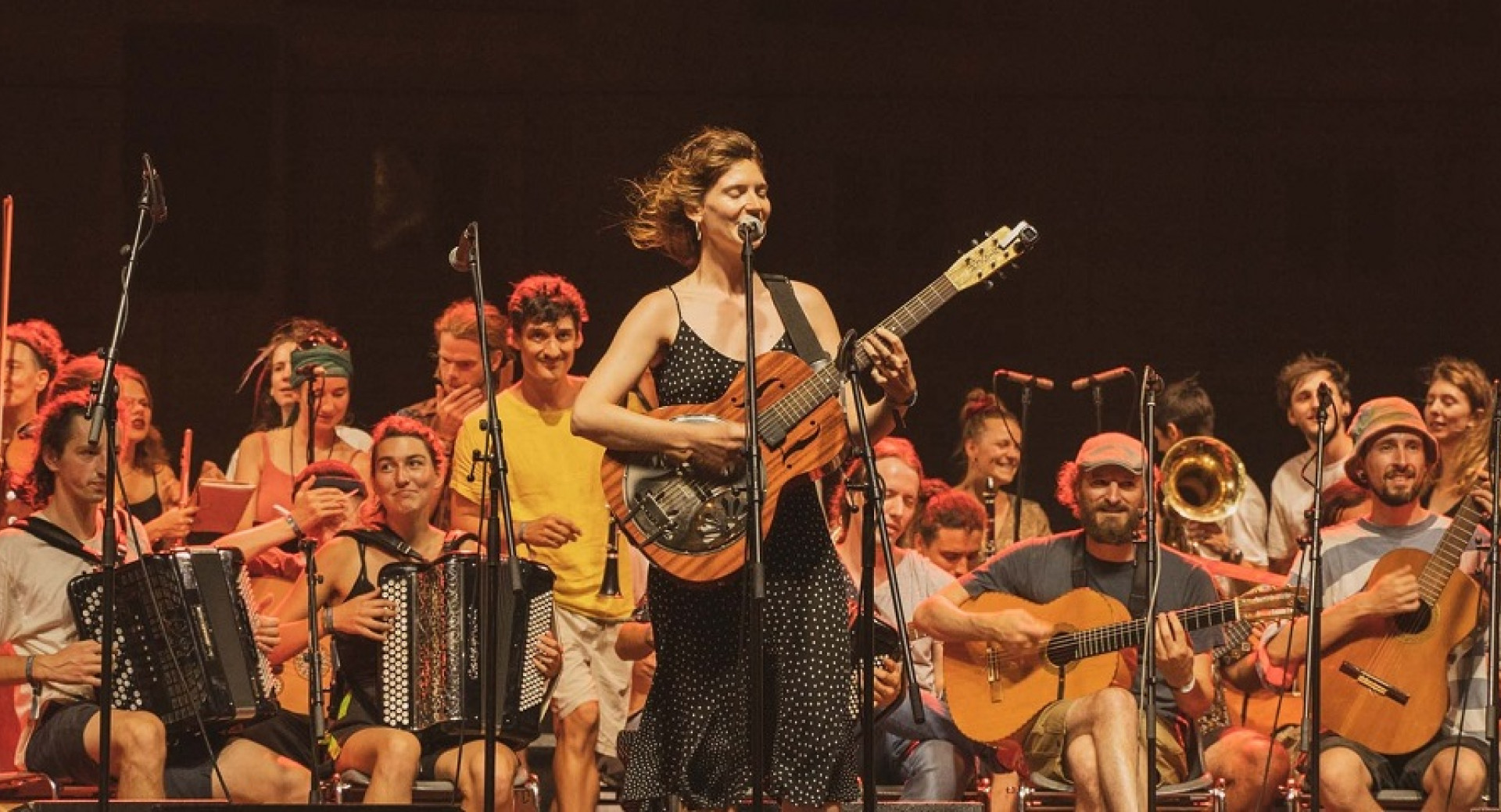 Etno orkester sedi in igra na instrumente, pred njimi pa stoji pevka s kitaro in poje v mikrofon.