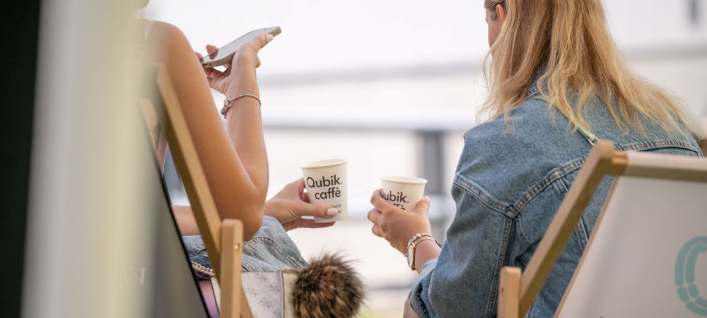 Two women, photographed from behind, are sitting on foldable wooden deck chairs, each holding a coffee in a paper cup.