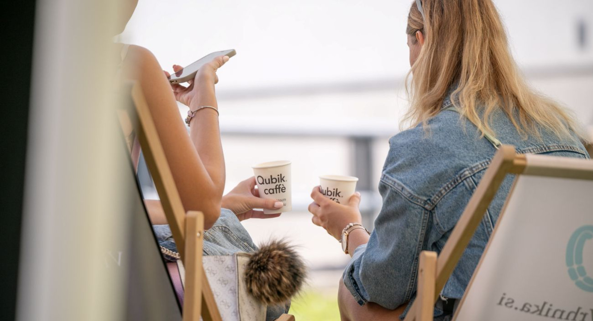 Two women, photographed from behind, are sitting on foldable wooden deck chairs, each holding a coffee in a paper cup.