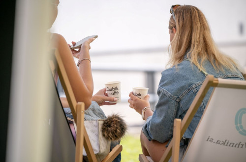 Two women, photographed from behind, are sitting on foldable wooden deck chairs, each holding a coffee in a paper cup.