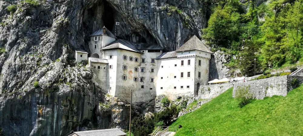 View from the bridge of a medieval castle built into a large rock cave, surrounded by high limestone cliffs and a green hillside in the foreground.