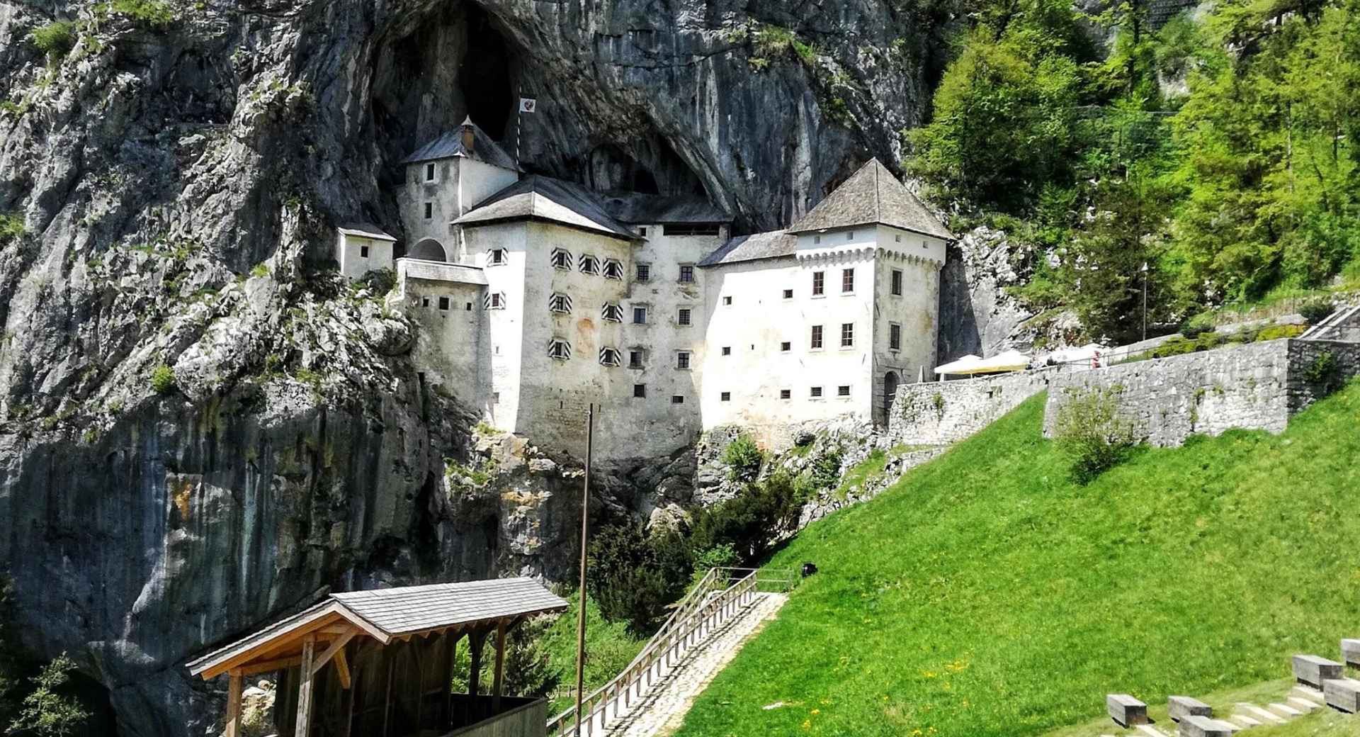 Blick von der Bruecke auf eine mittelalterliche Burg, die in eine grosse Felshoehle gebaut ist, umgeben von hohen Kalksteinfelsen und einem gruenen Huegel im Vordergrund.