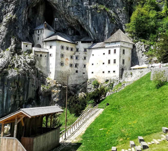 Blick von der Bruecke auf eine mittelalterliche Burg, die in eine grosse Felshoehle gebaut ist, umgeben von hohen Kalksteinfelsen und einem gruenen Huegel im Vordergrund.