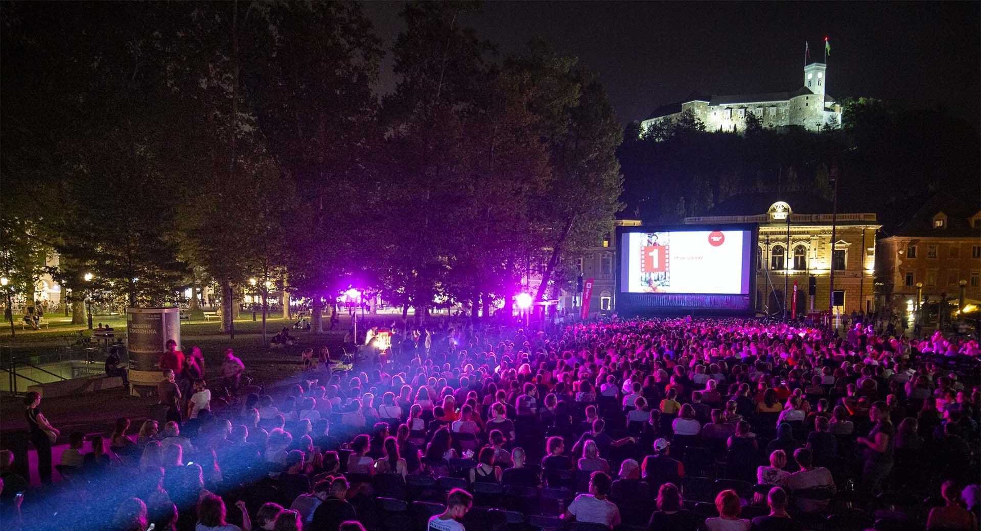 Visitors at an open-air cinema sit in the square at night, watching a film, with an illuminated castle in the background.