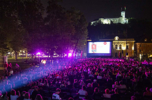 Visitors at an open-air cinema sit in the square at night, watching a film, with an illuminated castle in the background.