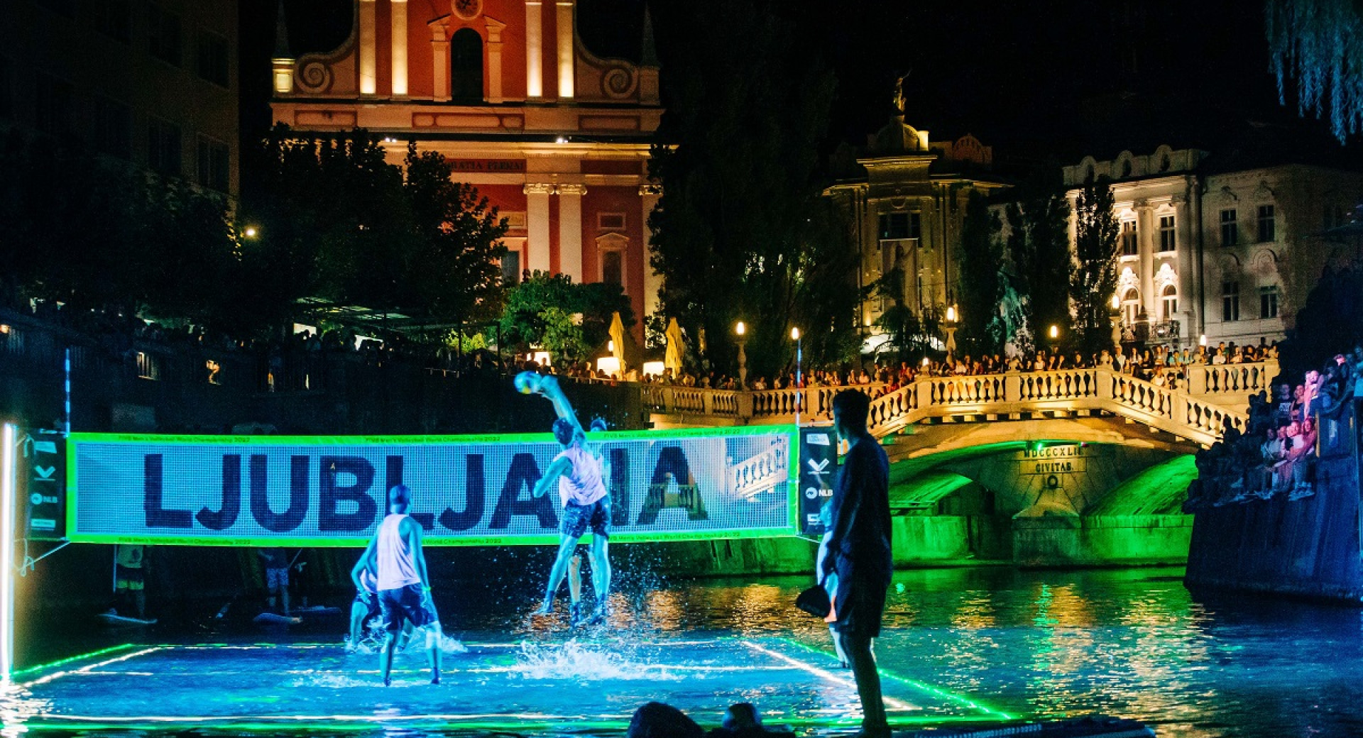 Men volleyball team plays on a floating field in a river. City lights in the background.