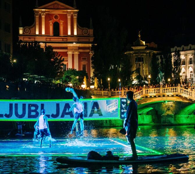 Men volleyball team plays on a floating field in a river. City lights in the background.