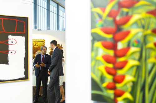 Dva moska v oblekah stojita v razstavnem prostoru. Okoli njiju razstavljene slikarske umetnine.