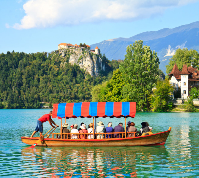 Tradicionalna lesena pletna z obiskovalci pluje po turkizno modri vodi Blejskega jezera, srednjevejski Blejski grad stoji na vrhu pecine, v ozadju pa so slikoviti Julijske Alpe.