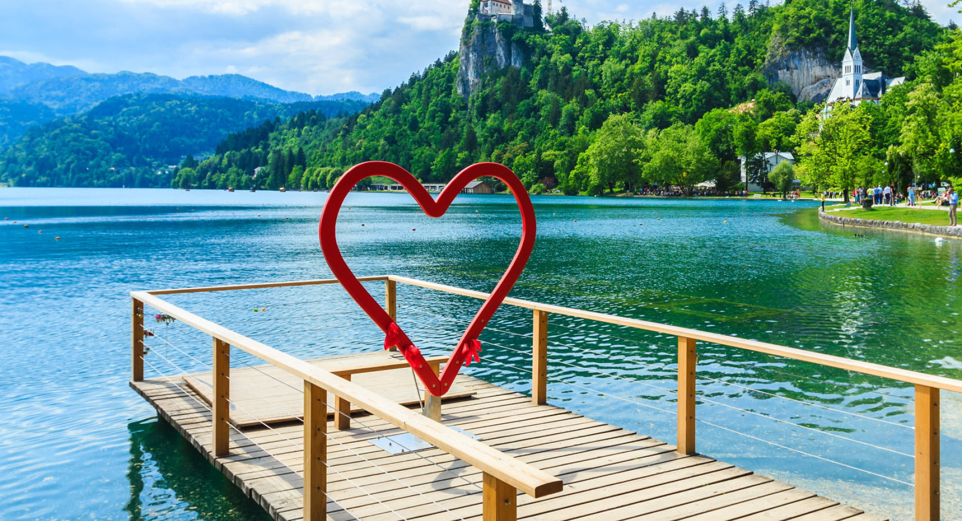 Lesen pomol z rdecim okvirjem v obliki srca stoji nad turkiznim jezerom, v ozadju pa so gozdnati hribi, cerkev in grad na skali.