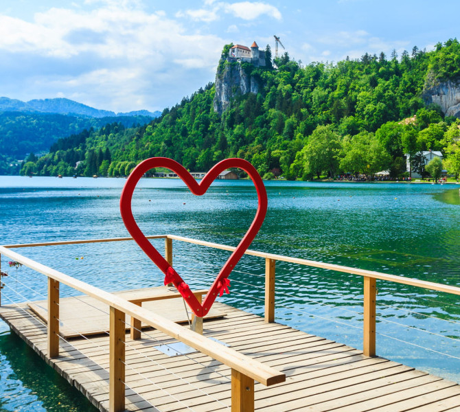 Ein Holzsteg mit einem roten herzförmigen Rahmen steht über einem türkisfarbenen See, im Hintergrund sind bewaldete Hügel, eine Kirche und eine Burg auf einem Felsen.