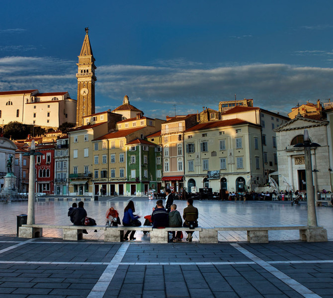 Una piazza circondata da edifici barocchi, con persone sedute sulle panchine in piumini.