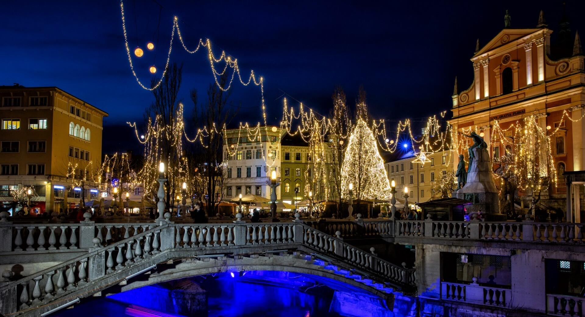 Boats on the river, with buildings and trees adorned with festive lights, glowing in vibrant colours in the evening.