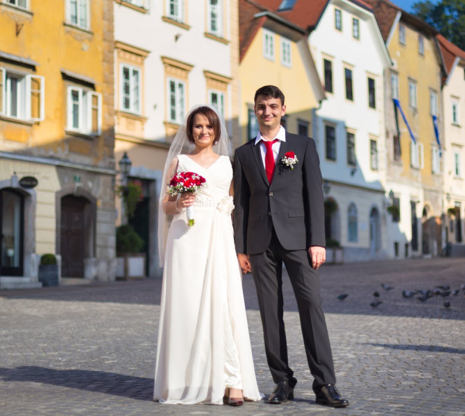 Nevesta v beli obleki s pajcolanom in sopkom rdecega cvetja ter zenin v crni obleki z belo srajco in rdeco kravato stojita v starem mestnem jedru.