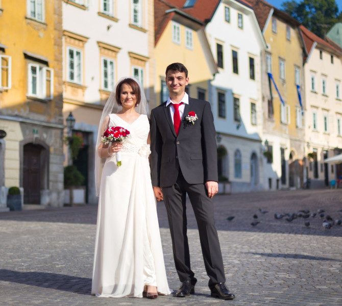 Nevesta v beli obleki s pajčolanom in šopkom rdečega cvetja in ženin v včrni obleki z belo srajco in rdečo kravato.