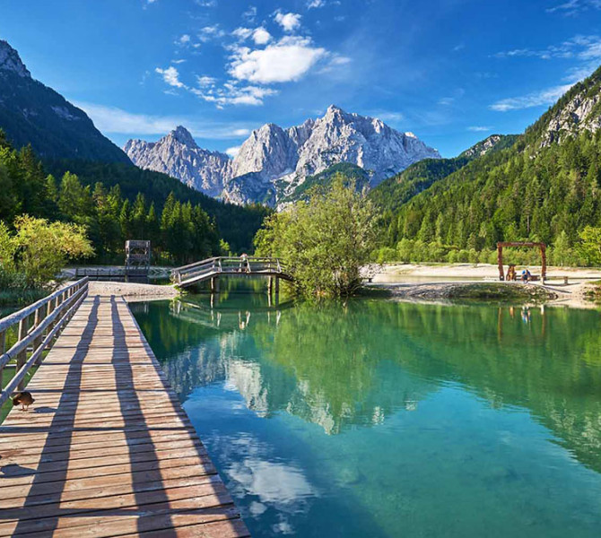 Un ponton en bois traverse la surface calme du lac Jasna vers des sommets montagneux pittoresques qui se reflètent dans l'eau turquoise sous un ciel bleu.