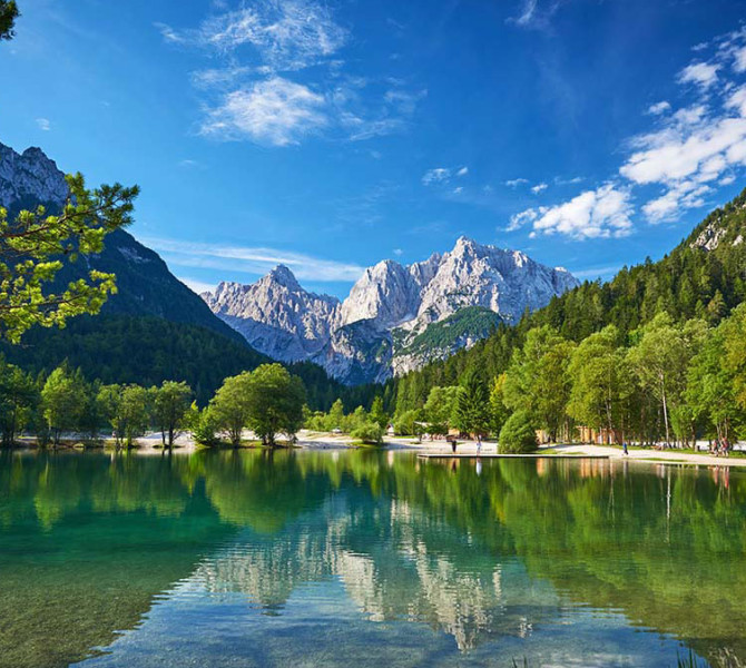 Une vue panoramique imprenable sur le lac calme de Jasna, reflétant les arbres verts et les hauts sommets rocheux des Alpes Juliennes sous un ciel bleu.