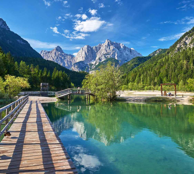 Ein Holzsteg führt über die ruhige Oberfläche des Jasna-Sees zu malerischen Berggipfeln, die sich im türkisfarbenen Wasser unter blauem Himmel spiegeln.