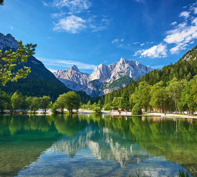 Čudovit panoramski pogled na mirno jezero Jasna, ki v svoji gladini zrcali zelena drevesa in visoke skalnate vrhove Julijskih Alp pod modrim nebom.