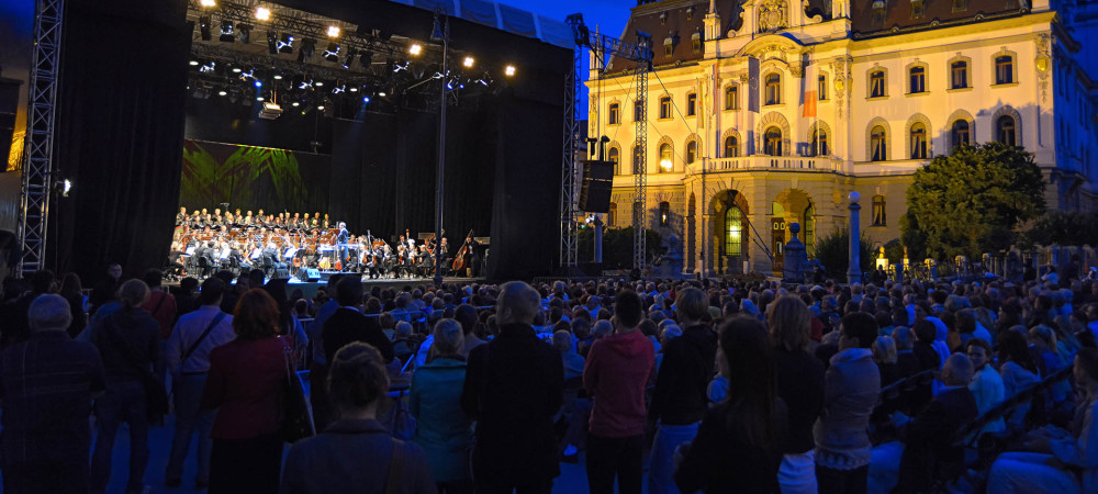 Orkester na odru, postavljenem na Kongresnem trgu, igra na festivalu Junij v Ljubljani, v ozadju pa se dviga osvetljena stavba Univerze v Ljubljani.