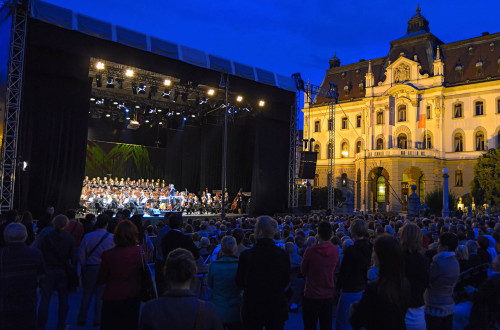 Orkester na odru, postavljenem na Kongresnem trgu, igra na festivalu Junij v Ljubljani, v ozadju pa se dviga osvetljena stavba Univerze v Ljubljani.