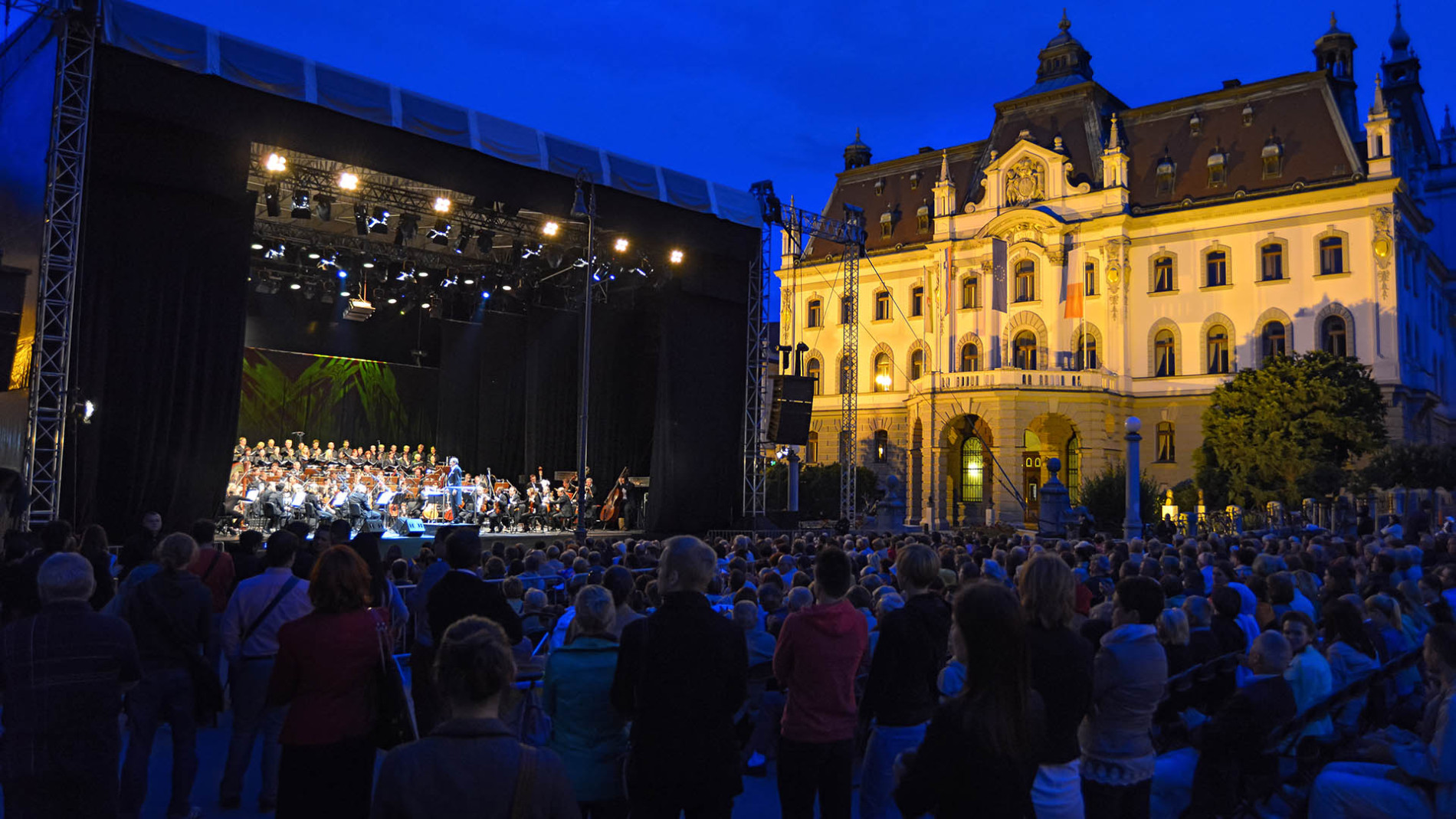 Orkester na odru, postavljenem na Kongresnem trgu, igra na festivalu Junij v Ljubljani, v ozadju pa se dviga osvetljena stavba Univerze v Ljubljani.