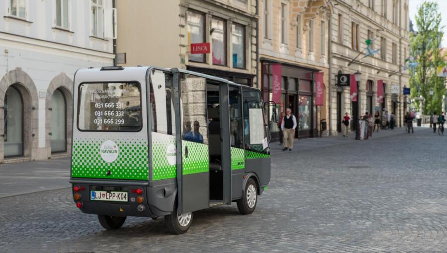 Un petit bus au design vert et blanc est garé sur une rue pavée, avec ses portes ouvertes. Des personnes passent devant des magasins le long de la rue.