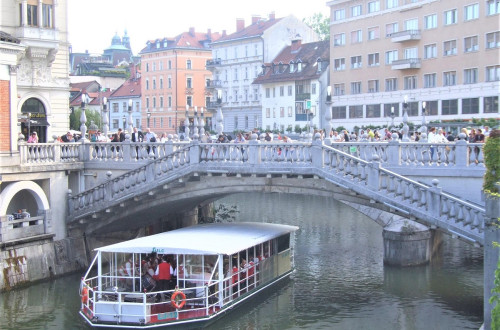 Na ladjici na reki Ljubljanici igra pihalna godba, ljudje pa jo z mostu opazujejo in poslusajo.